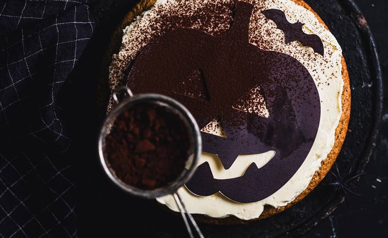 En Halloween-kake med et gresskaransikt som blir dekorert med melis, sett ovenfra.