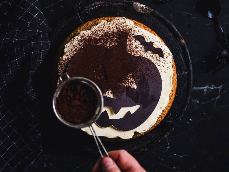 En Halloween-kake med et gresskaransikt som blir dekorert med melis, sett ovenfra.