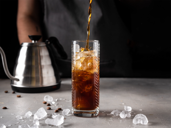 En person heller iskald kaffe fra en termos i et høyt glass med isbiter, omgitt av isbiter på bordet.