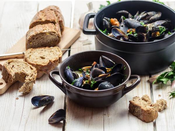 Skål med blåskjell i en svart gryte, omgitt av brød og noen skjell på et lyst bord.