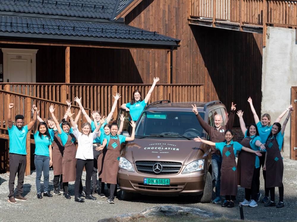 En gruppe jenter står foran en bil med armene i været, foran en trehus med treverk og en terrasse.