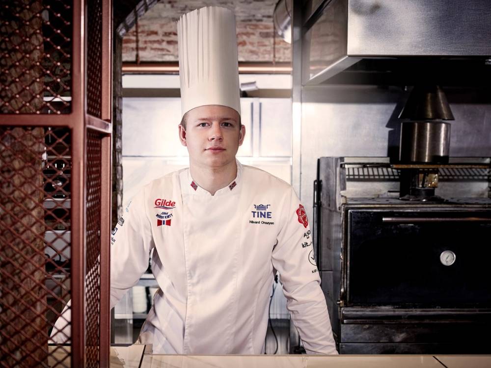 En kok står bak en kjøkkenøy i et profesjonelt kjøkken, iført hvit kokkeuniform med navn og logoer.