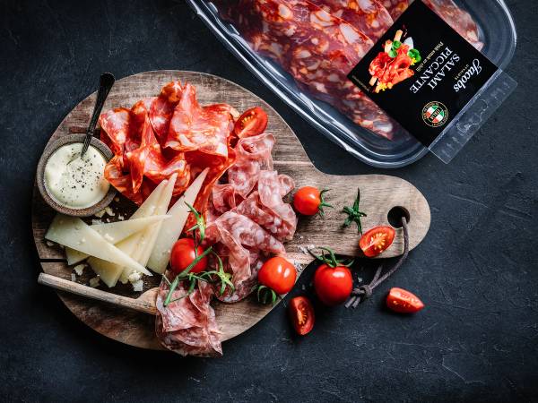 Spekemat med ulike typer skinke og kjøtt, cherrytomater og ost på et rundt trefat, med en pakke med spekemat i bakgrunnen.