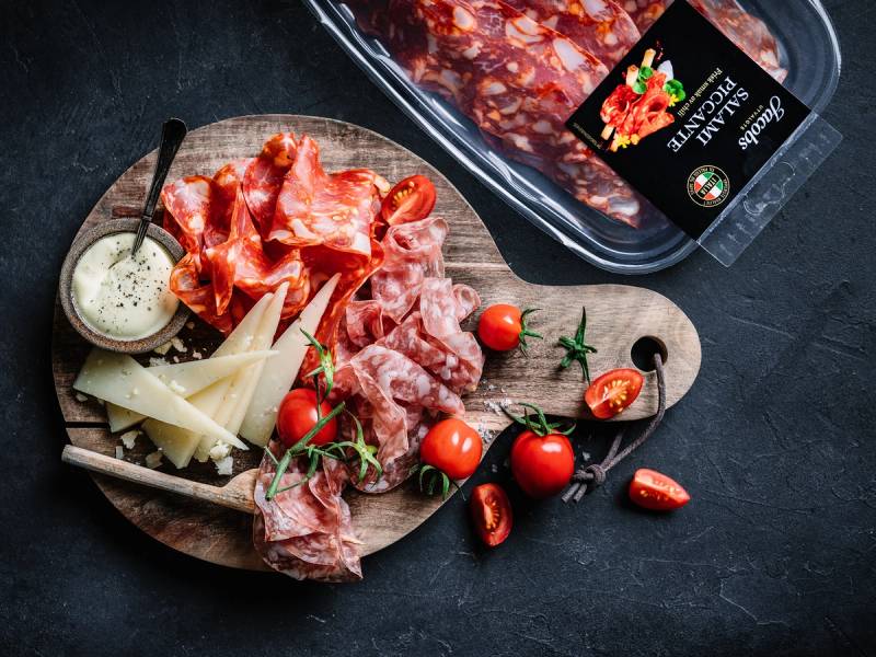 Spekemat med ulike typer skinke og kjøtt, cherrytomater og ost på et rundt trefat, med en pakke med spekemat i bakgrunnen.