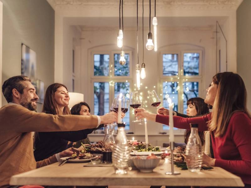 En gruppe mennesker som skåler med vin rundt et spisebord i et lyst rom med store vinduer og hengende lys.