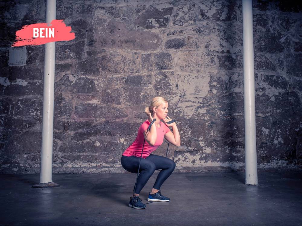 En person i rosa treningsklær gjør en knebøy foran en murvegg, med stangstativ på hver side.