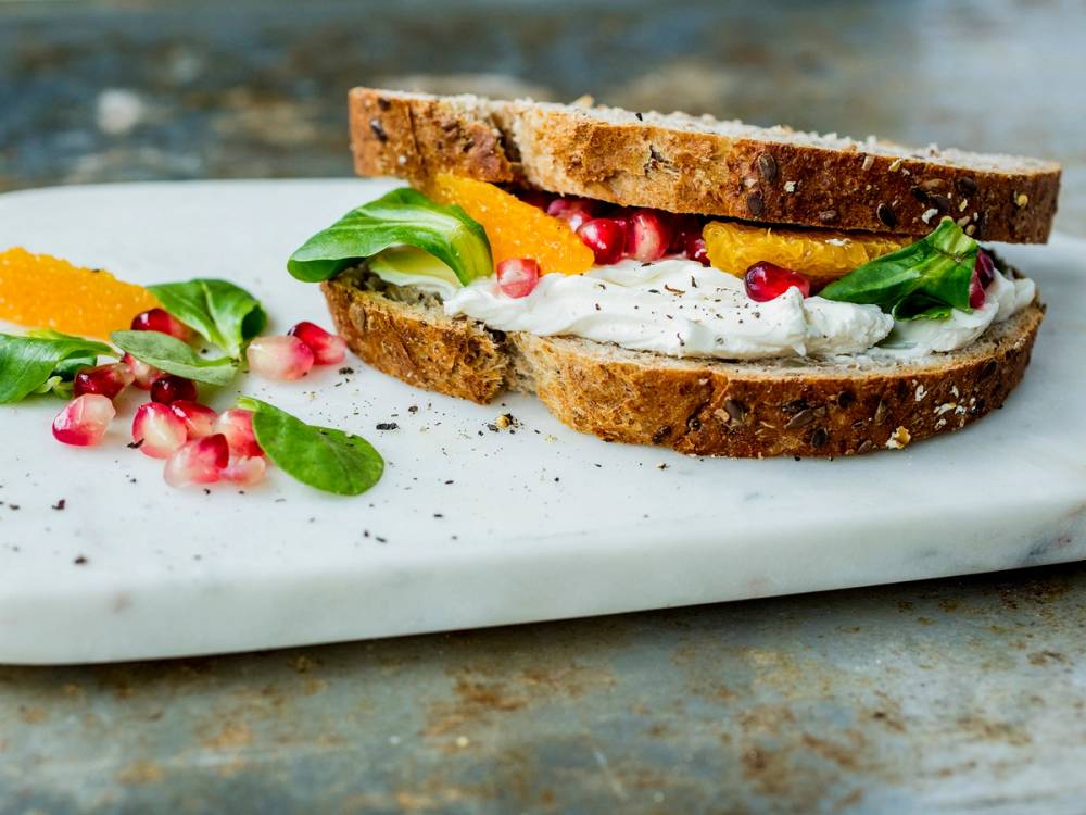 Mozzarellaost på en skive grovt brød med grønne urter og granateplefrø, ser ut som en enkel sandwich eller snack.