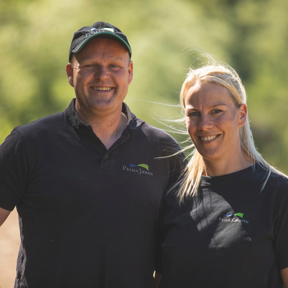To personer står utendørs med grønne trær i bakgrunnen, begge har på seg svarte t-skjorter med logo, og ansiktene er utvisket.