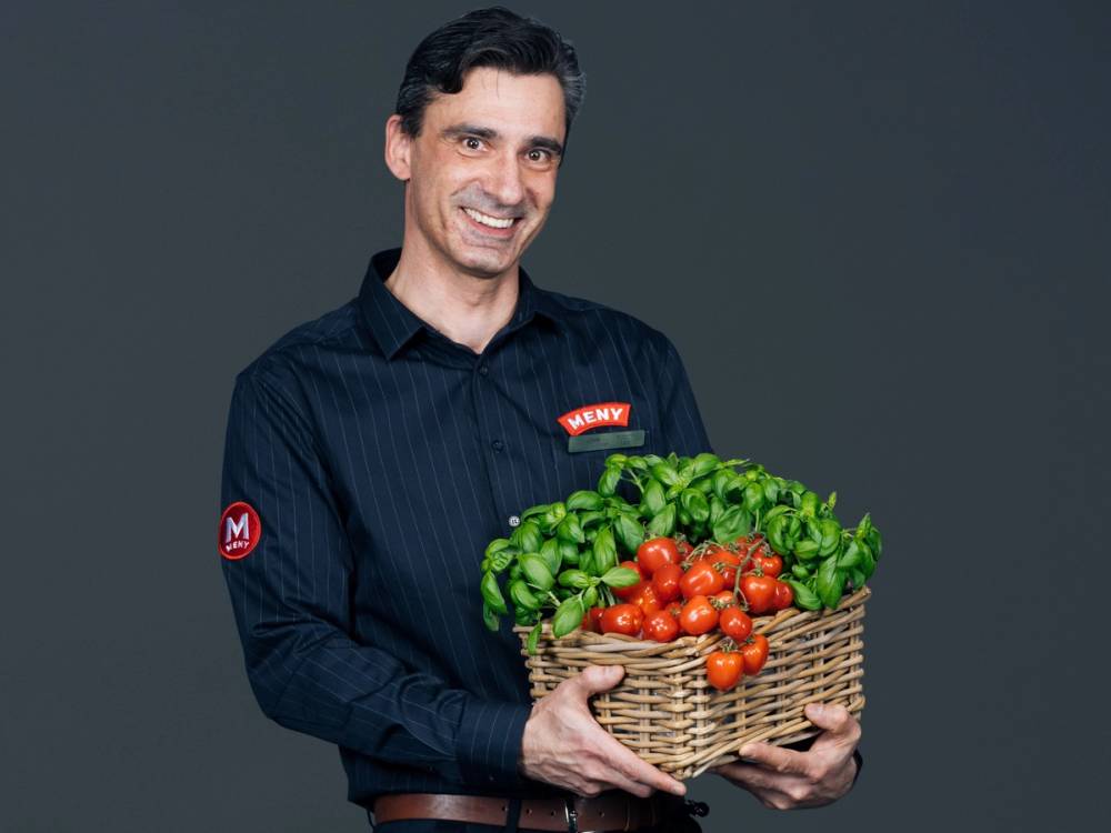 Person holder en kurv med tomat og frisk basilikum mot en grå bakgrunn.