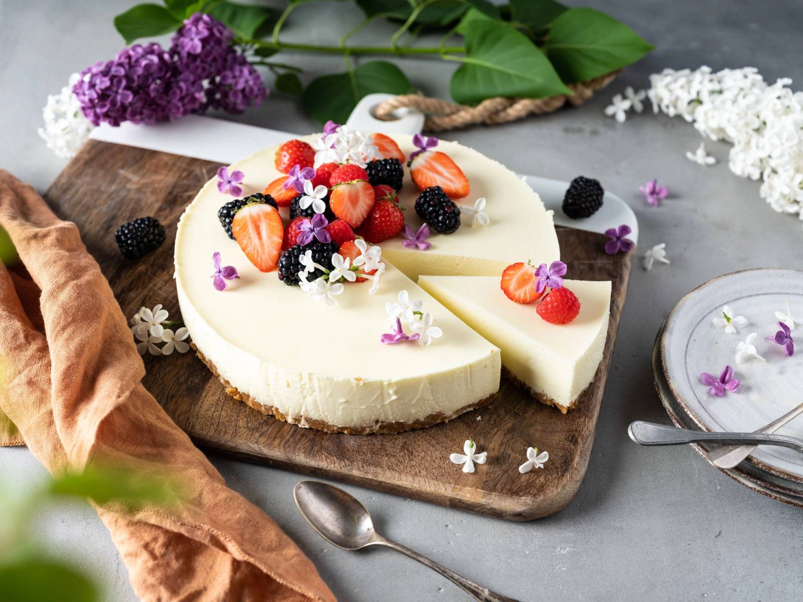 En rund festkake med hvit glasur pyntet med ferske jordbær, bringebær og små spiselige blomster, plassert på et trebrett med blomster rundt.
