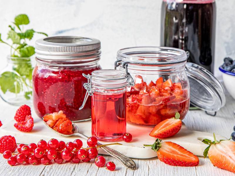 Syltetøy og saft i glasskrukker omgitt av jordbær og rips på et hvitt bord.