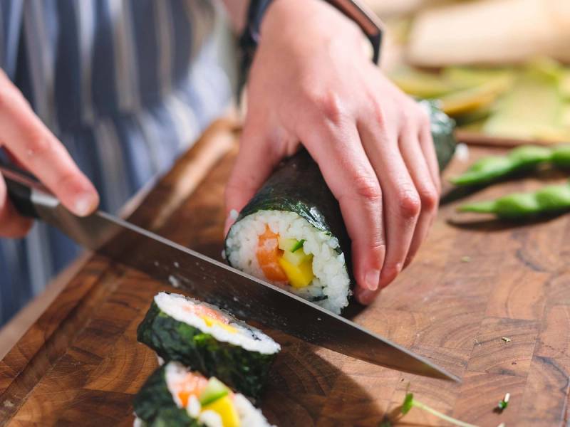 Person skjærer opp en rull med sushi på et trebord, med flere andre sushi-ruller rundt.