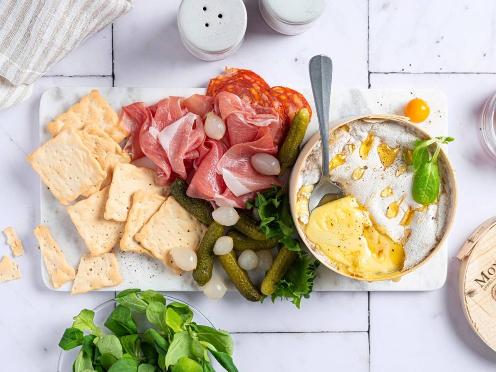 Et fat med spekeskinke, syltetøy, grønnsaker og en liten bolle med egg og urter på et hvitt bord.