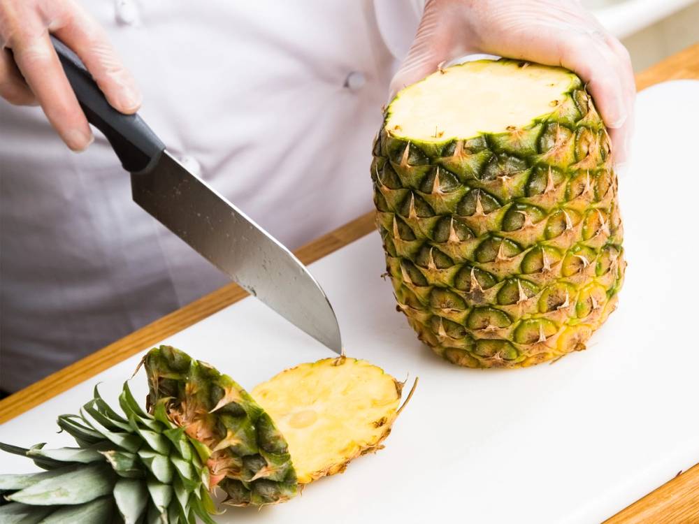 En person skjærer av toppen av en ananas med en kniv på kjøkkenet.
