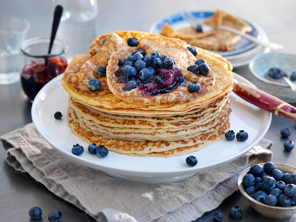 Pannekaker med blåbær og lønnesirup på en hvit tallerken, omgitt av friske blåbær og en skje med syltetøy.