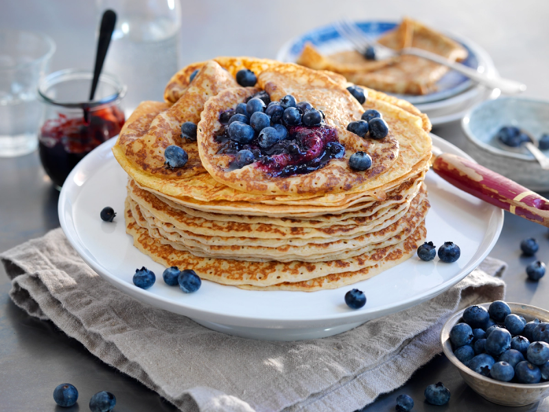 Pannekaker med blåbær og lønnesirup på en hvit tallerken, omgitt av friske blåbær og en skje med syltetøy.