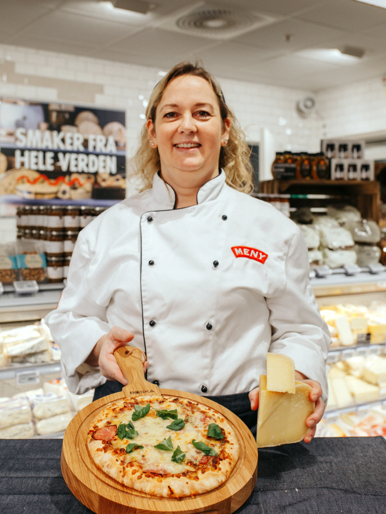 En ansatt i MENY-kjøkkenuniform holder en pizza og et stykke ost i en butikk med ost og matvarer i bakgrunnen.