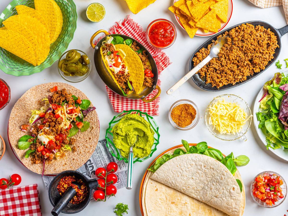 Tacos med kjøtt, grønnsaker og ost på en tallerken, omgitt av tilbehør som guacamole, salsa og tortillachips, på et bord med fargerike elementer.
