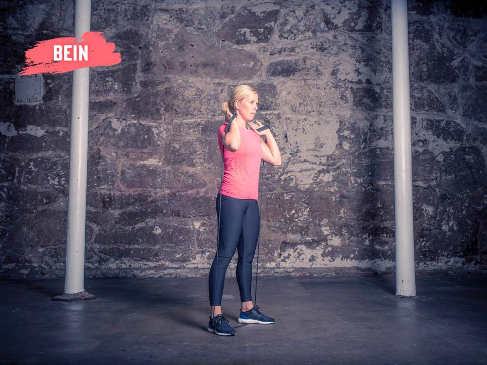 En kvinne utfører en knebøy med strikk rundt skuldrene i et treningsrom med en murvegg i bakgrunnen.