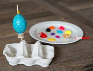 Et egg er malt blå med gullfargede prikker og står i en eggekartong, med en malepensel og en palett med ulike farger av maling i nærheten.