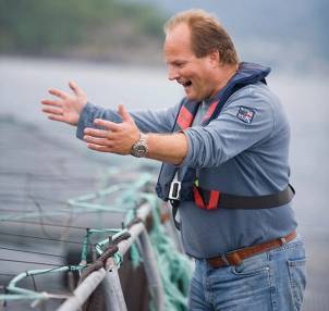 En mann med blå jakke og armbåndsur står ved en kai og viser noe til noen utenfor bildet, med vann og natur i bakgrunnen.