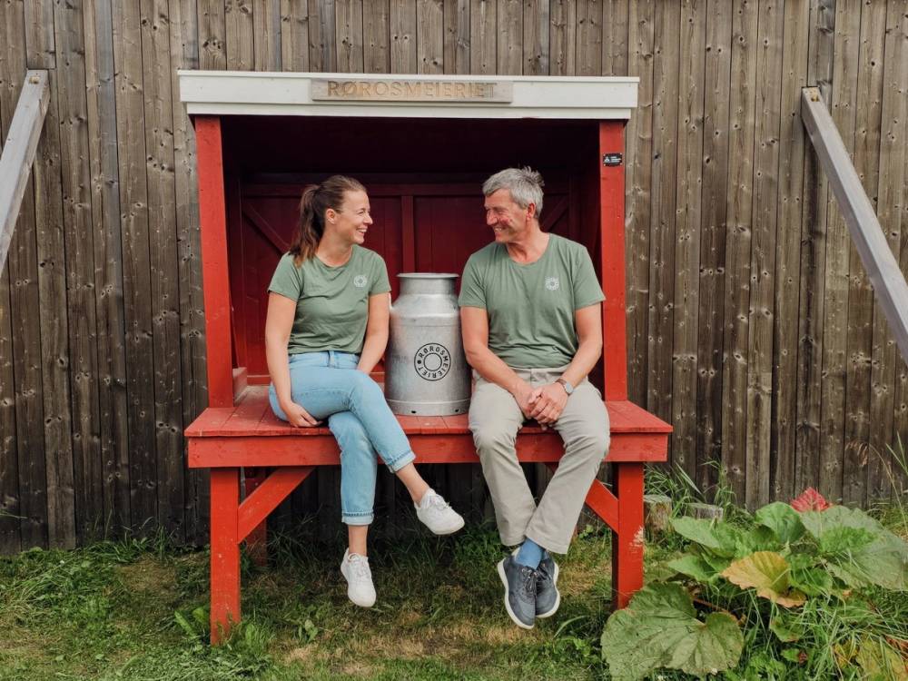 To personer sitter på en benk foran en melkekanne, med en trevegg i bakgrunnen og blomster i forgrunnen.
