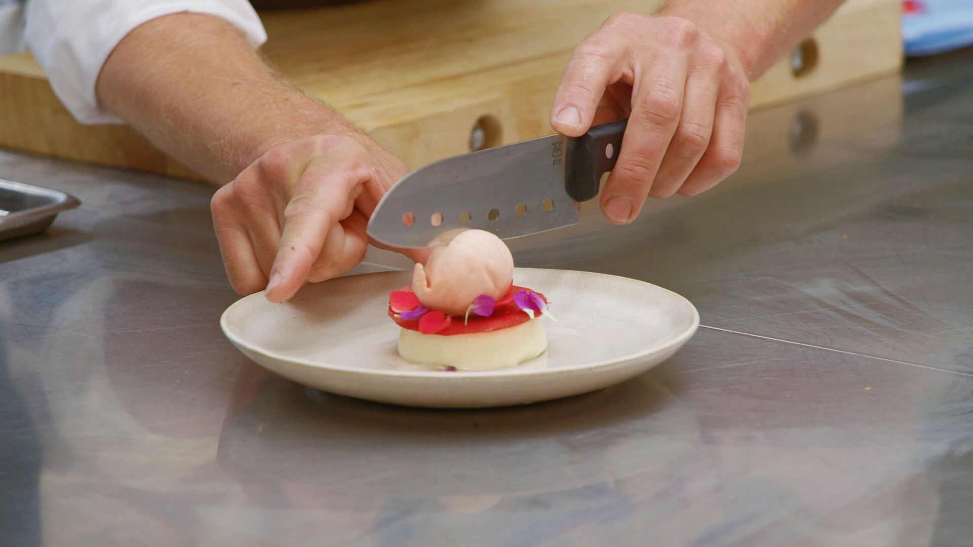En person skjærer opp en rund, rosa dessert med en kniv på et hvitt fat, med hendene synlige i bildet.