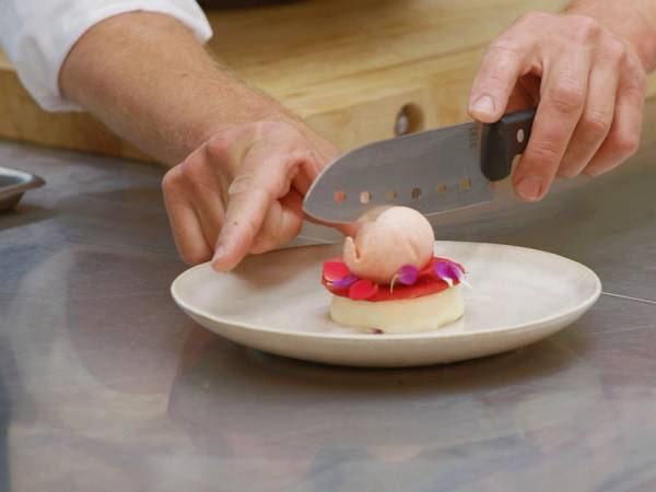 En person skjærer opp en rund, rosa dessert med en kniv på et hvitt fat, med hendene synlige i bildet.