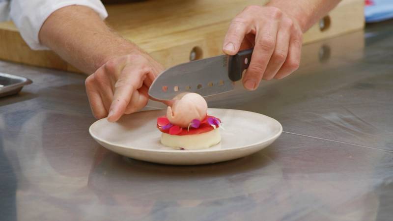 En person skjærer opp en rund, rosa dessert med en kniv på et hvitt fat, med hendene synlige i bildet.