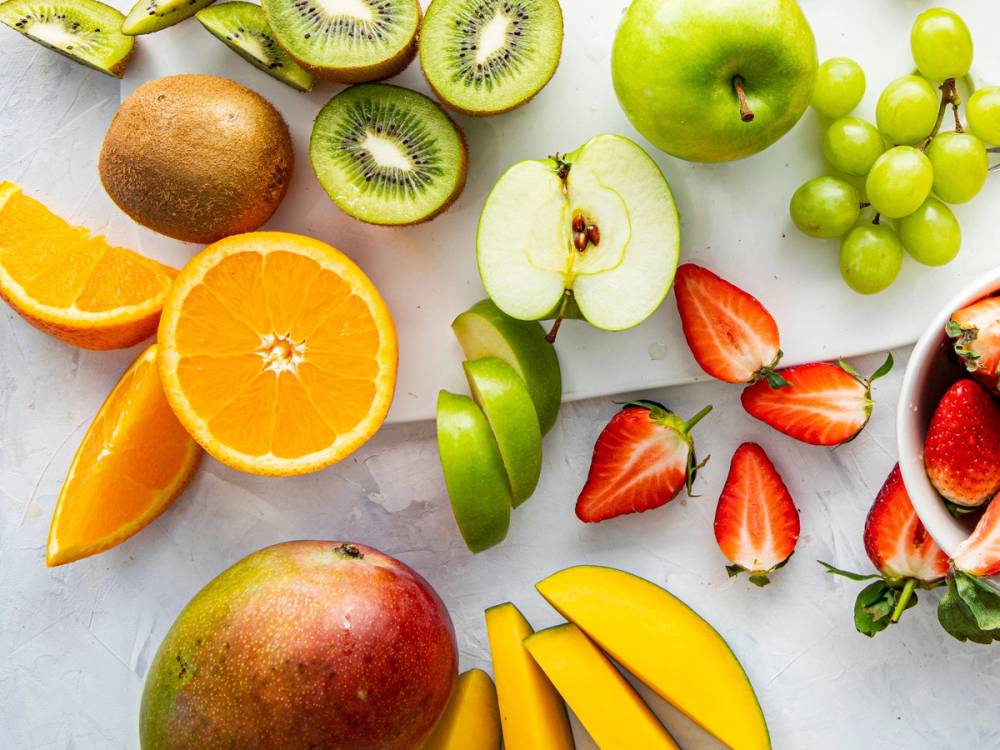 Ulike frukter som kiwi, eple, appelsin, jordbær, druer og mango er kuttet opp og lagt ut på en hvit overflate.