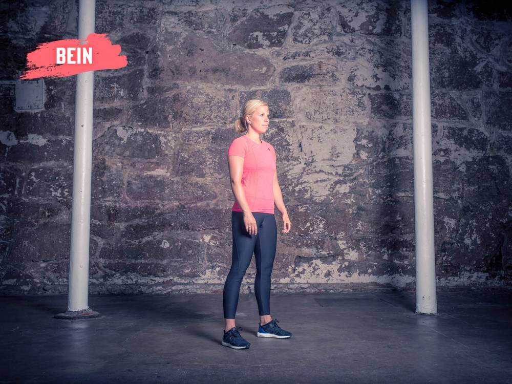En kvinne står i en treningshall med en grå murvegg og to stenger på hver side, iført rosa t-skjorte og mørke treningsbukser.