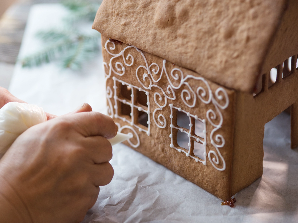 En person pynter et pepperkakehus med hvit glasur, med et ferdig dekorert pepperkakehus i fokus.