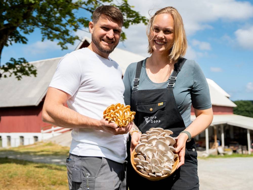 To personer viser frem sopper de har plukket, med en bygning og blå himmel i bakgrunnen.