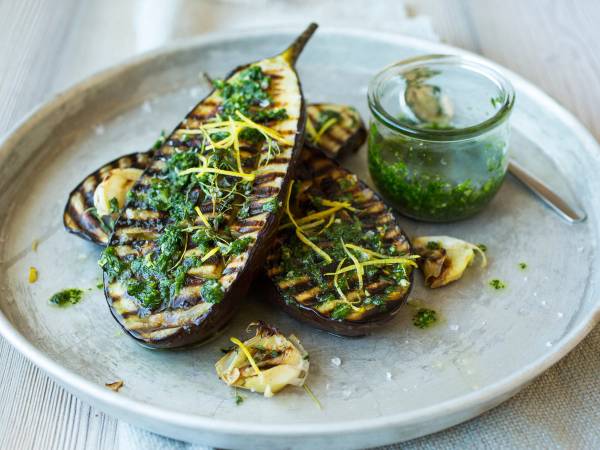 Grillet aubergine med frisk grønn urtesaus og sitronzest på en tallerken, med en liten glasskål med saus ved siden av.