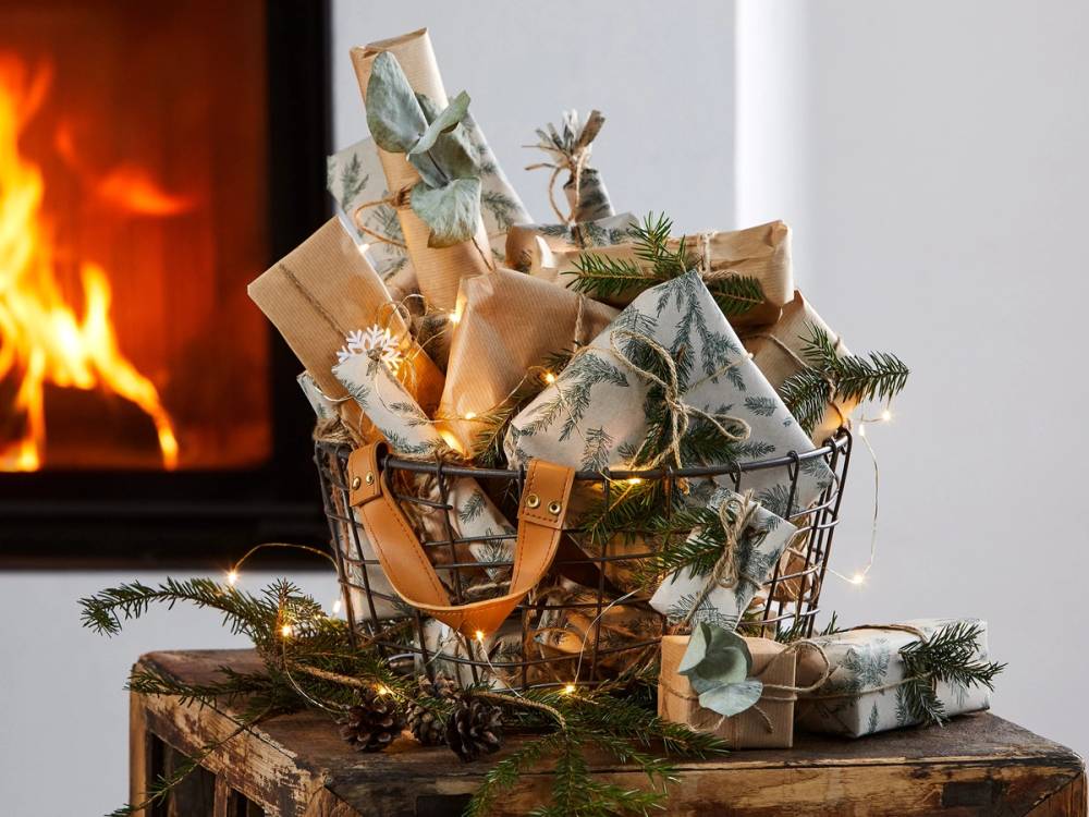 En julehandelkurv fylt med gaver og julepynt står på et bord foran en peis, dekorert med granbar og julelys.