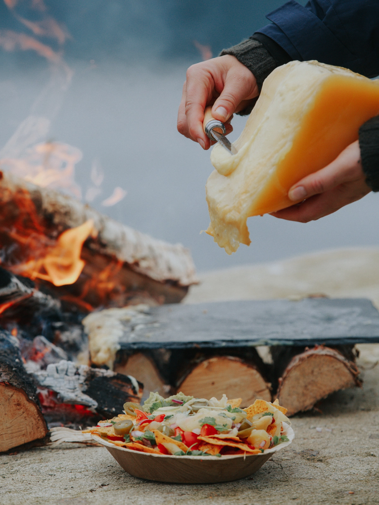 Person holder en stor skive alpeost over en grill med flammer, med en tallerken med pasta og grønnsaker foran.