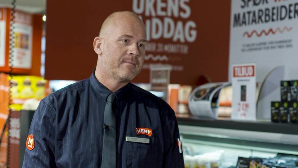 En person i uniform står bak disken på en matbutikk, med produkter og reklameskilt i bakgrunnen.