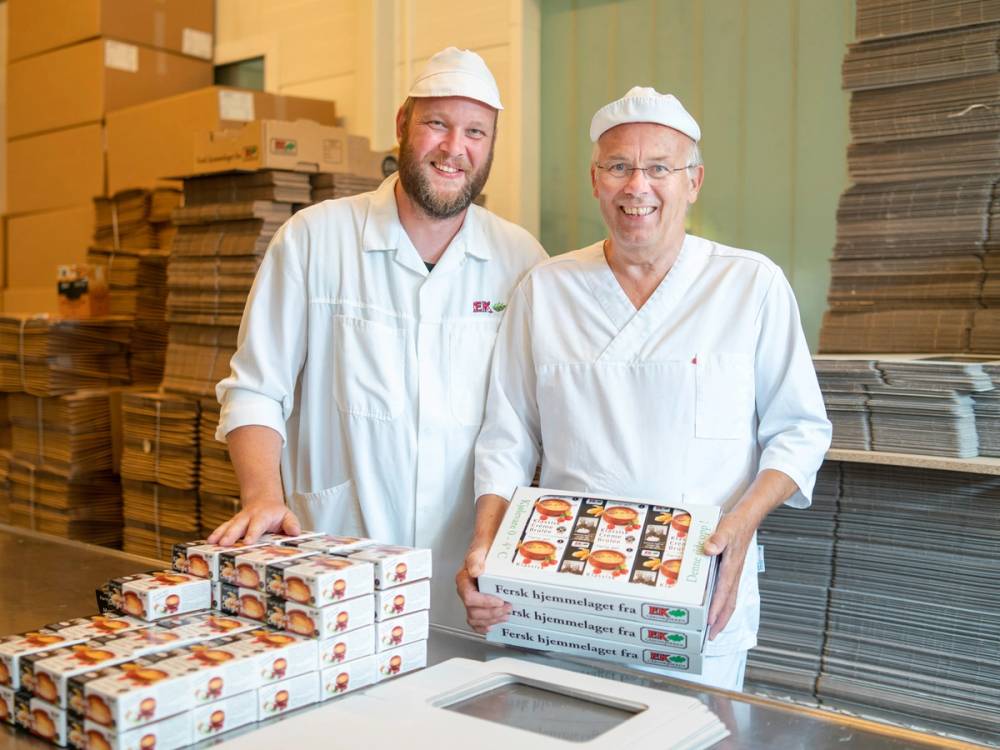 To personer i arbeidsklær står i en pappemballasje-fabrikk med stabler av ferdige pizzaer og pappesker rundt seg.