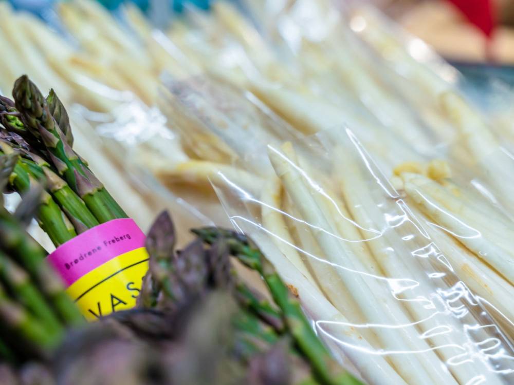 Ferske asparges pakket i plast på en butikkhylle, med en rosa og gul etikett i nærheten.