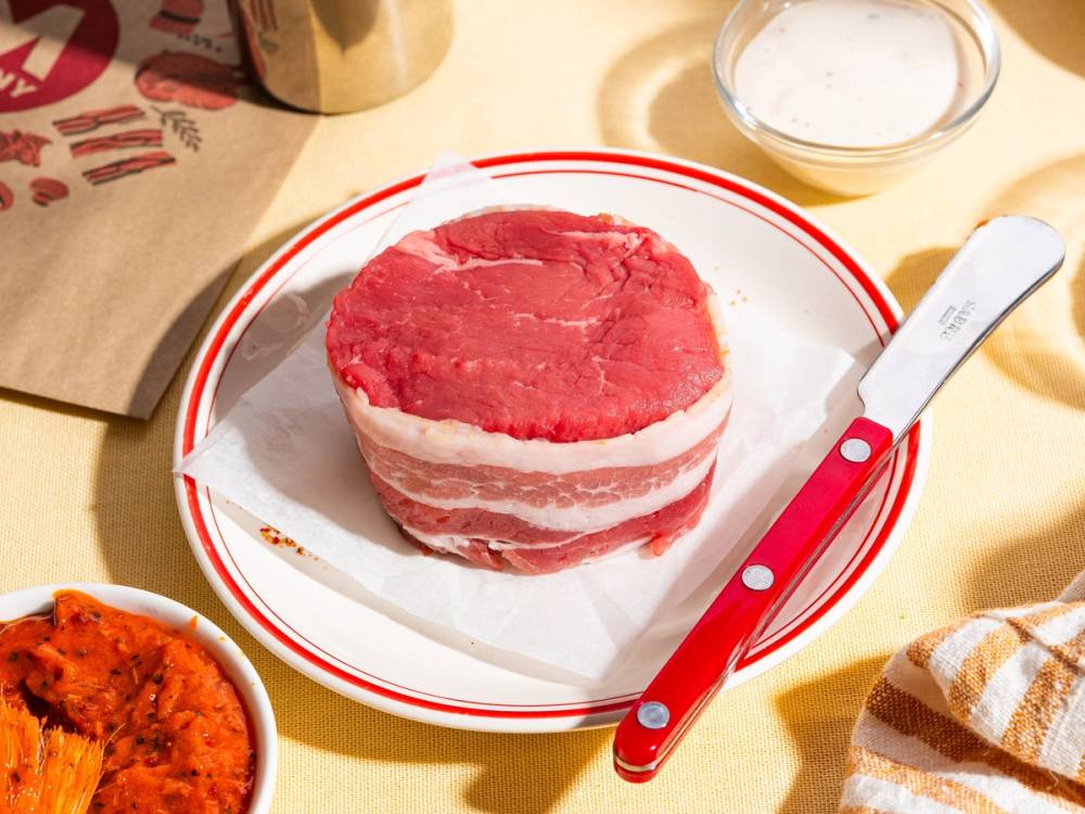 En skive av ferskt kjøtt med innpakket bacon på en tallerken, omgitt av ulike matvarer og kjøkkenutstyr.