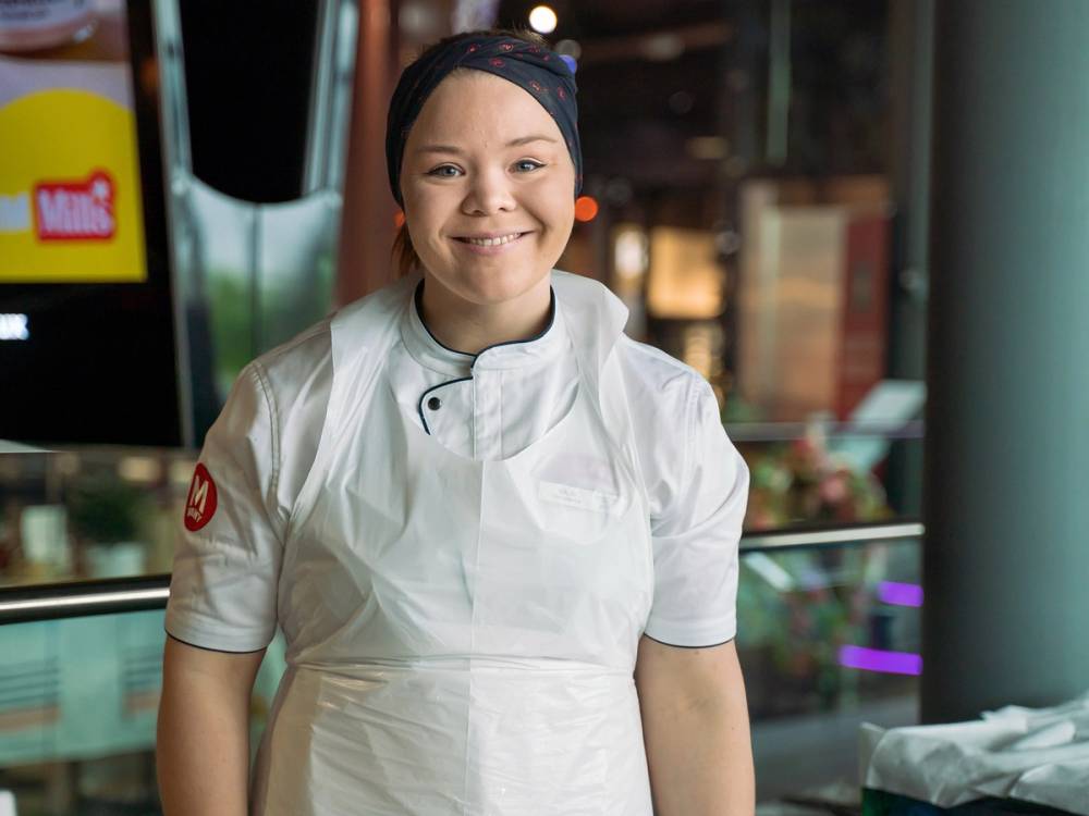 En person i hvit kokkeforkledning står foran en butikk eller restaurant, med en butikkdør og vindu i bakgrunnen.