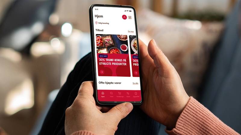 Person som holder en smarttelefon med en app som viser tilbud på matvarer fra MENY, sett fra siden.