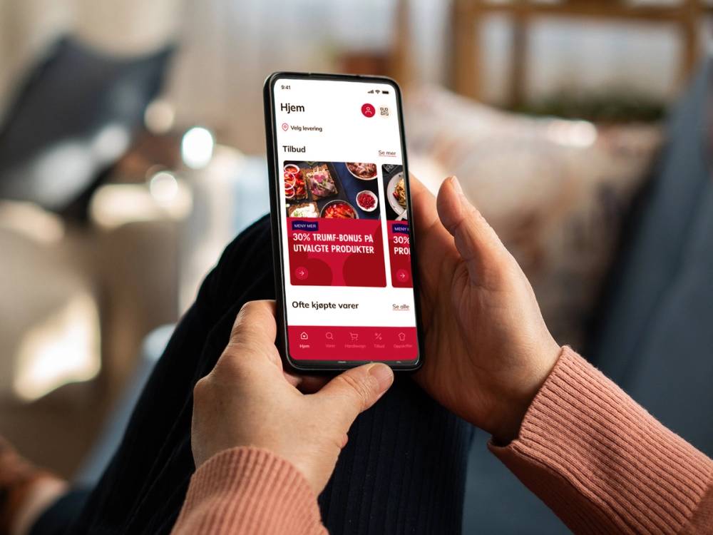 Person som holder en smarttelefon med en app som viser tilbud på matvarer fra MENY, sett fra siden.