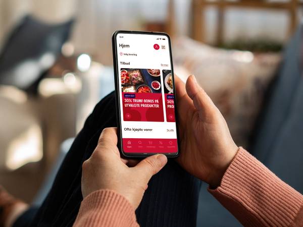Person som holder en smarttelefon med en app som viser tilbud på matvarer fra MENY, sett fra siden.