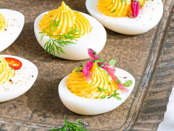 Små påskeegg fylt med gul krem og dekorert med rosa og grønne spiselige blomster og blader, plassert på en treplate.