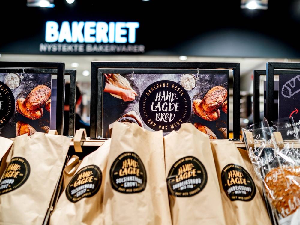 Utsalgsdisk med brød og bakevarer på en matbutikk, med en digital skjerm i bakgrunnen som reklamerer for håndlagde brød.