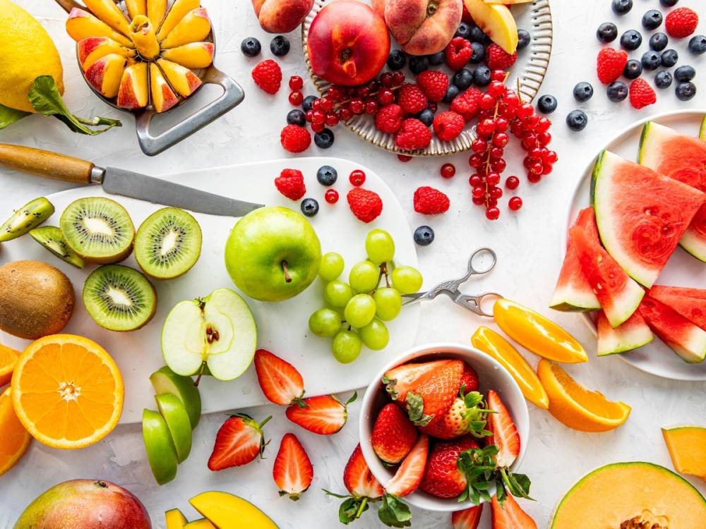 Fargerike oppskårne frukter som vannmelon, kiwi, eple, jordbær, blåbær og druer er lagt ut på et bord, med noen kjøkkenredskaper rundt.