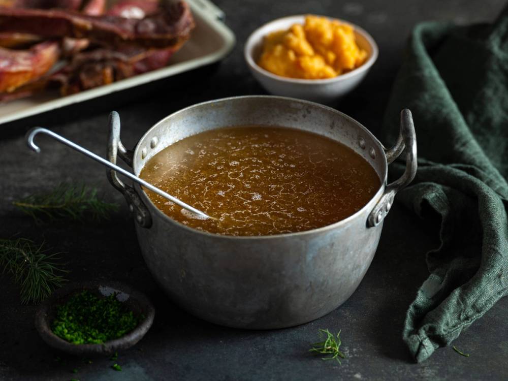 En metallkjele med kraftig sjy og en skje oppi, omgitt av tilbehør som potetmos og grønnsaker, på et mørkt bord.