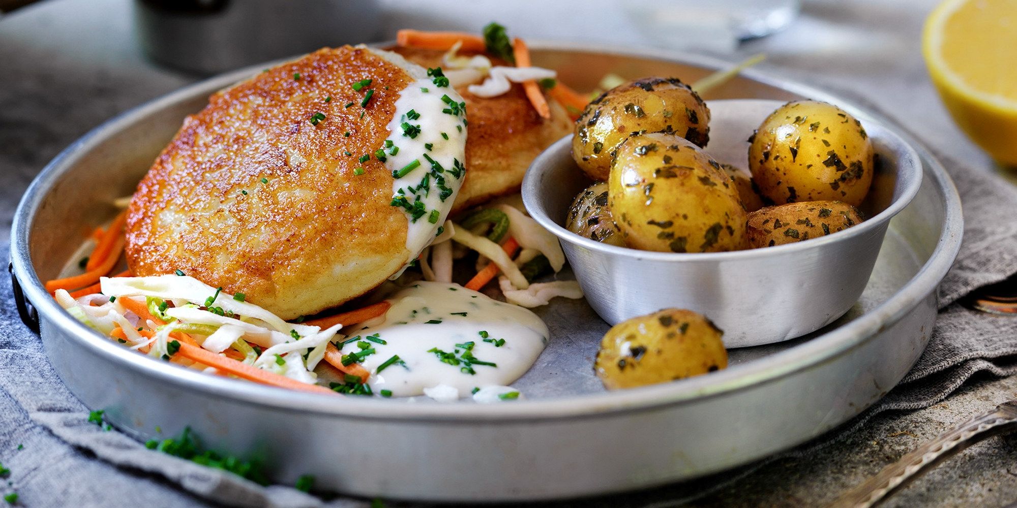 Fiskekaker med råkostsalat og små kokte poteter på en tallerken, med sitron i bakgrunnen.