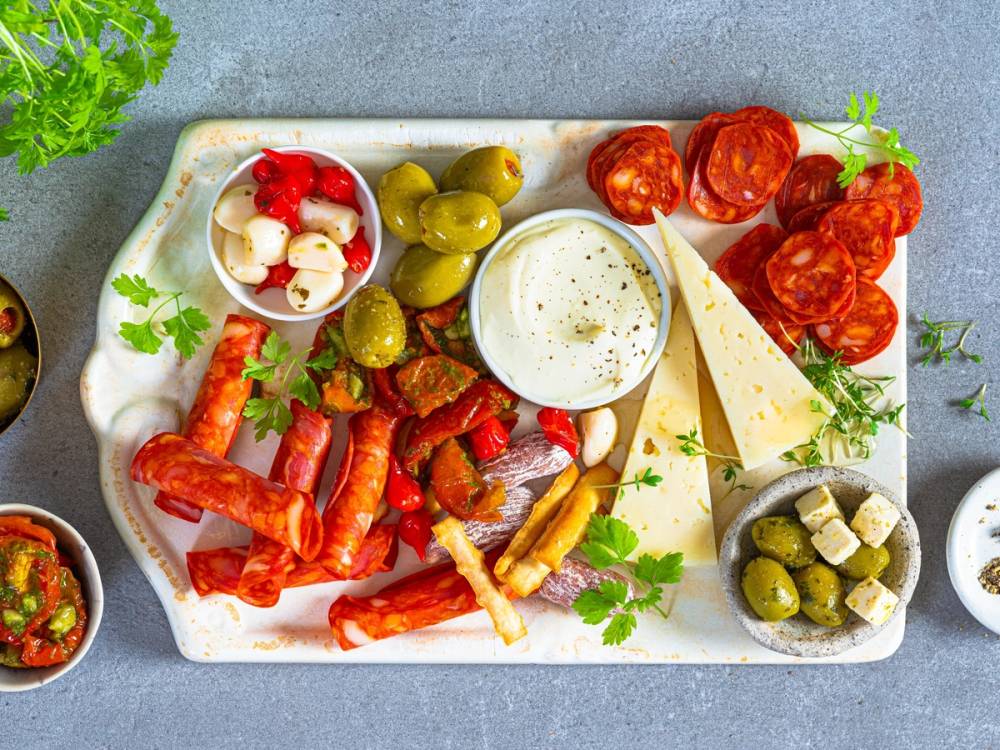 Et fat med ulike tapasretter, inkludert oliven, cherrytomater, ost, og spekepølser, på en lys bakgrunn.